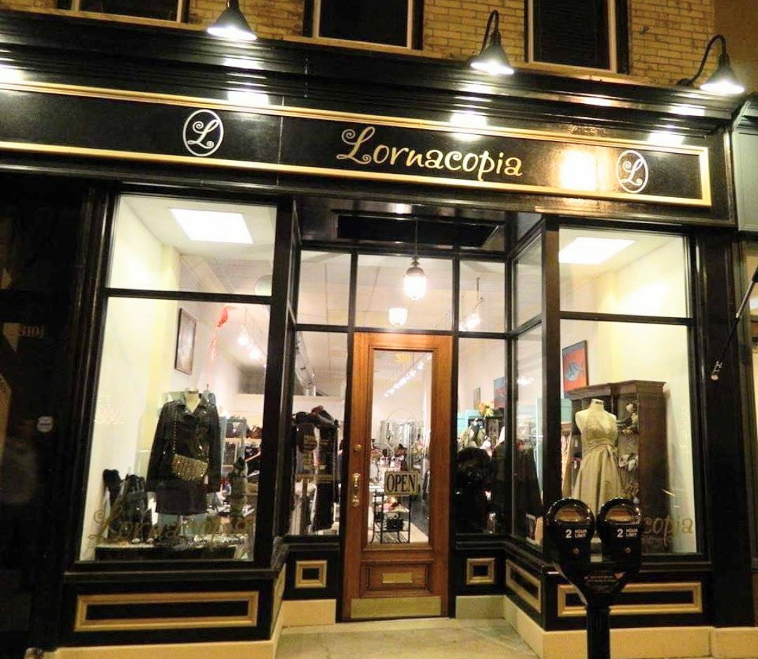 Lornacopia boutique storefront at night with black trim, gold lettering, large display windows, a wooden door, and mannequins visible inside the shop.